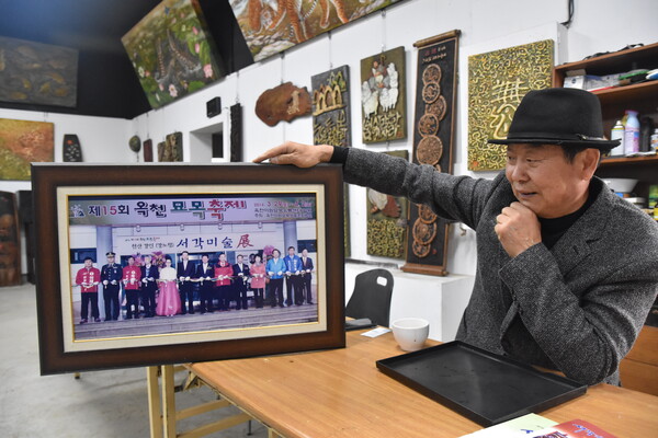 2014년 3월 옥천묘목축제 때 그가 서각미술전을 열어 기념촬영을 했던 사진을 보여주고 있다. 